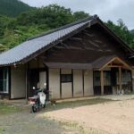 古民家の外観。瓦屋根と土壁の伝統的な日本家屋が緑の山に囲まれて建っている。
