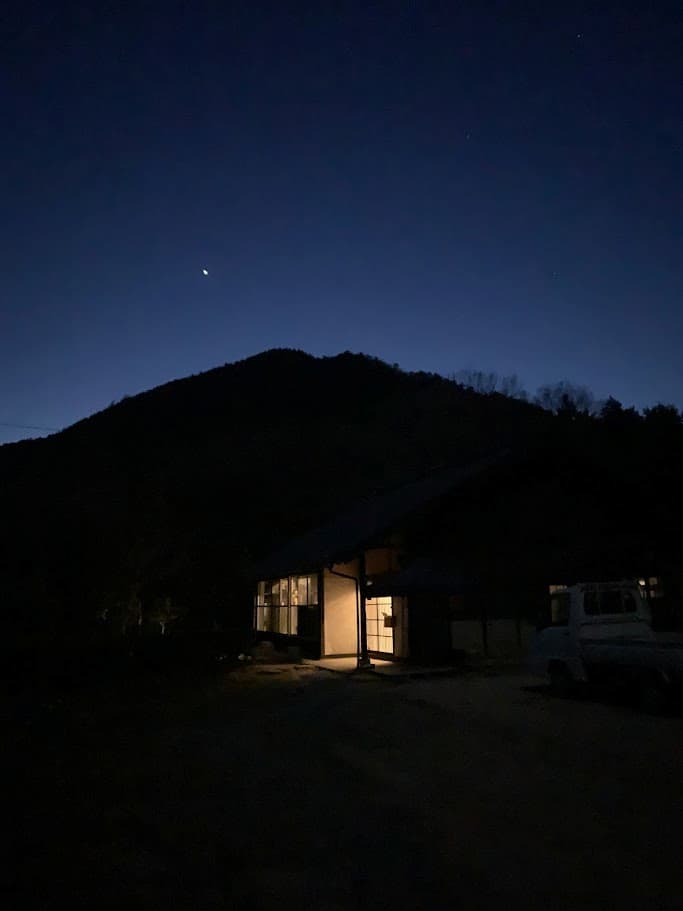 阿東の古民家の夜景。山のシルエットと星空の下、室内の灯りが温かく輝いている。