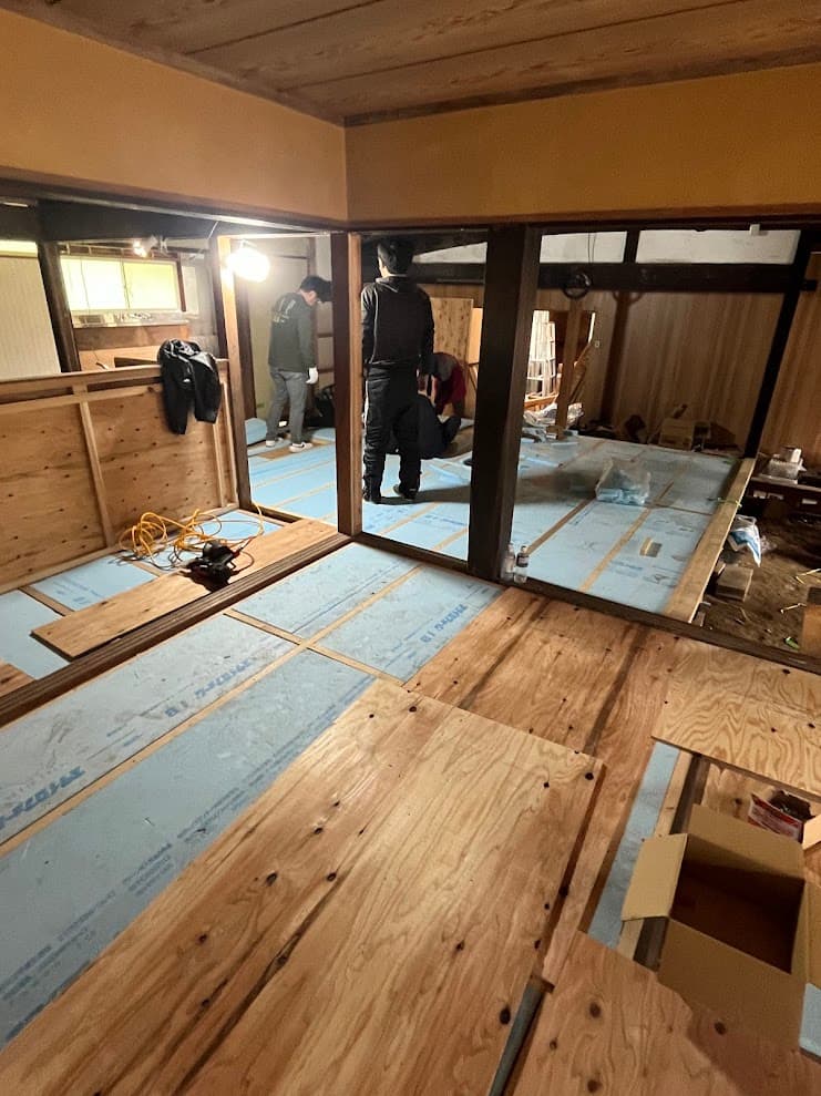 断熱材を敷き終えた床の上で、複数人が合板を張る作業を行っている古民家改修現場。