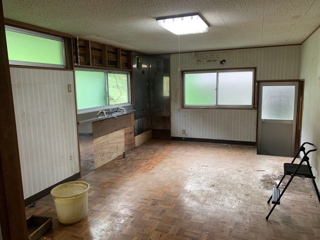改修前の古いキッチン。壁や床が傷み、流し台が撤去された状態で全体に古びた印象が残る。