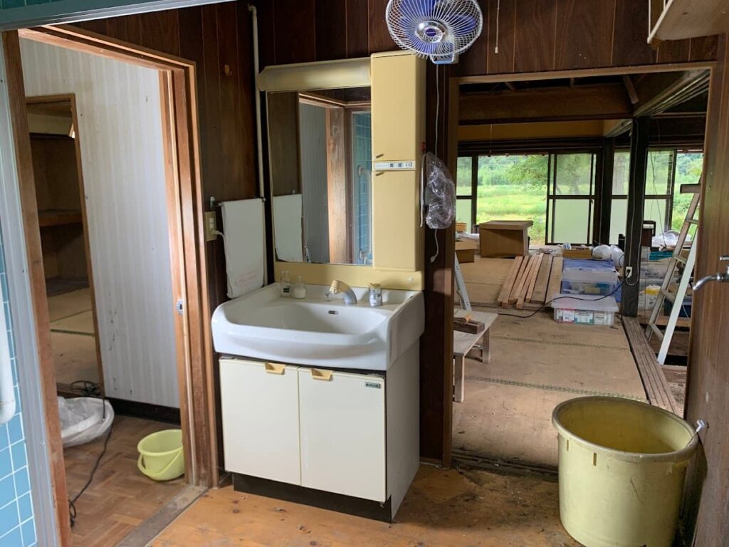 改修前の洗面所。木壁と古い洗面台が残り、経年による劣化が目立つ状態。奥には居間が見える。