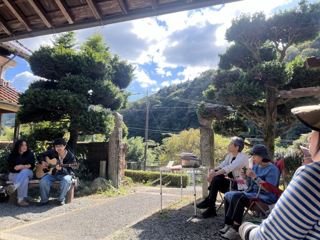 古民家の庭先で、ギター演奏を楽しむ演者と、椅子に座って聴いている参加者たちの様子。