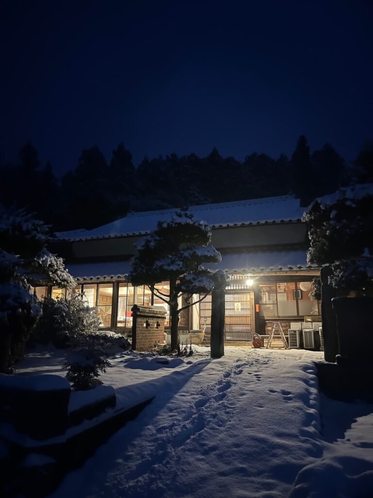 雪に包まれた夜のかんぬしの宿外観。灯りがともる古民家の玄関。